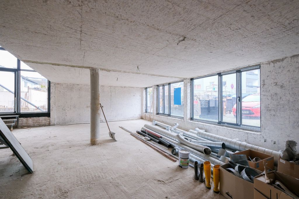 Ein leerer, unfertiger Raum mit Betonwänden und -decke, großen Fenstern, einem Stützpfeiler in der Raummitte und Rohren sowie Baumaterialien auf dem Boden.