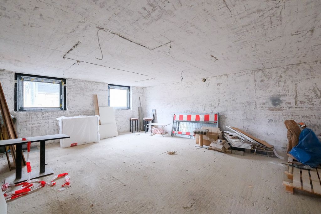 Leerer, unfertiger Kellerraum mit Sichtbetonwänden, zwei kleinen Fenstern und verstreut gelagertem Baumaterial.