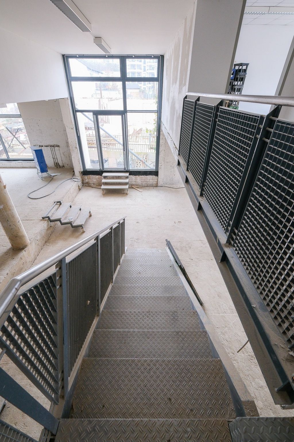 Blick von oben auf eine Metalltreppe mit Gittern als Geländer in einem unvollendeten oder renovierungsbedürftigen Raum mit großen Fenstern.