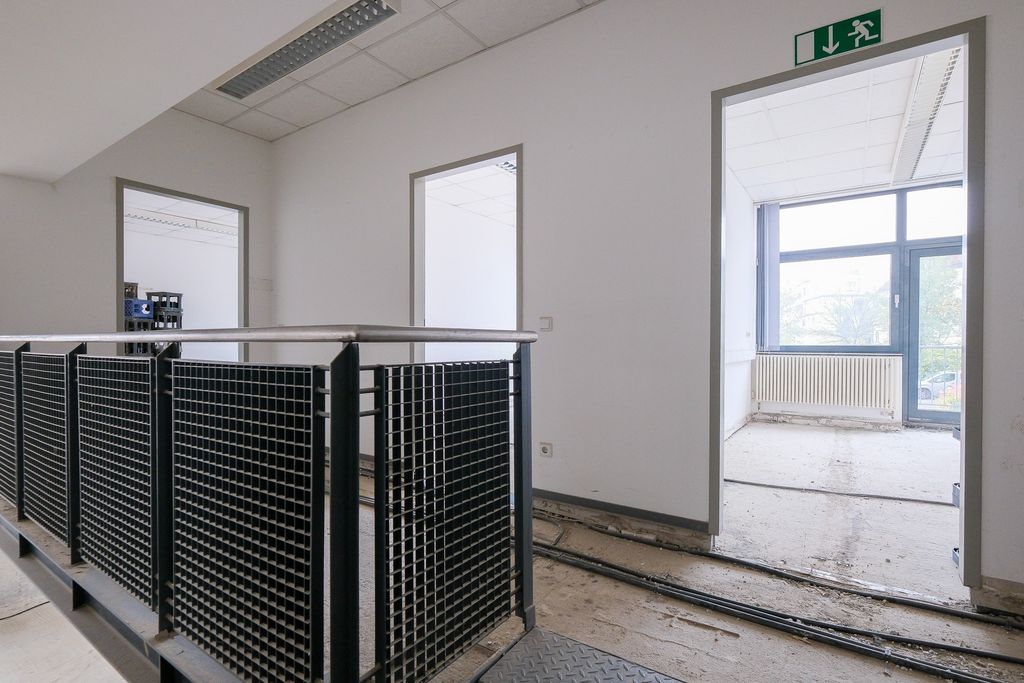 Flur in einem unmöblierten Bürogebäude mit grauen Metallgeländern, weißen Wänden, zwei Türen und großen Fenstern mit Sicht auf Bäume.