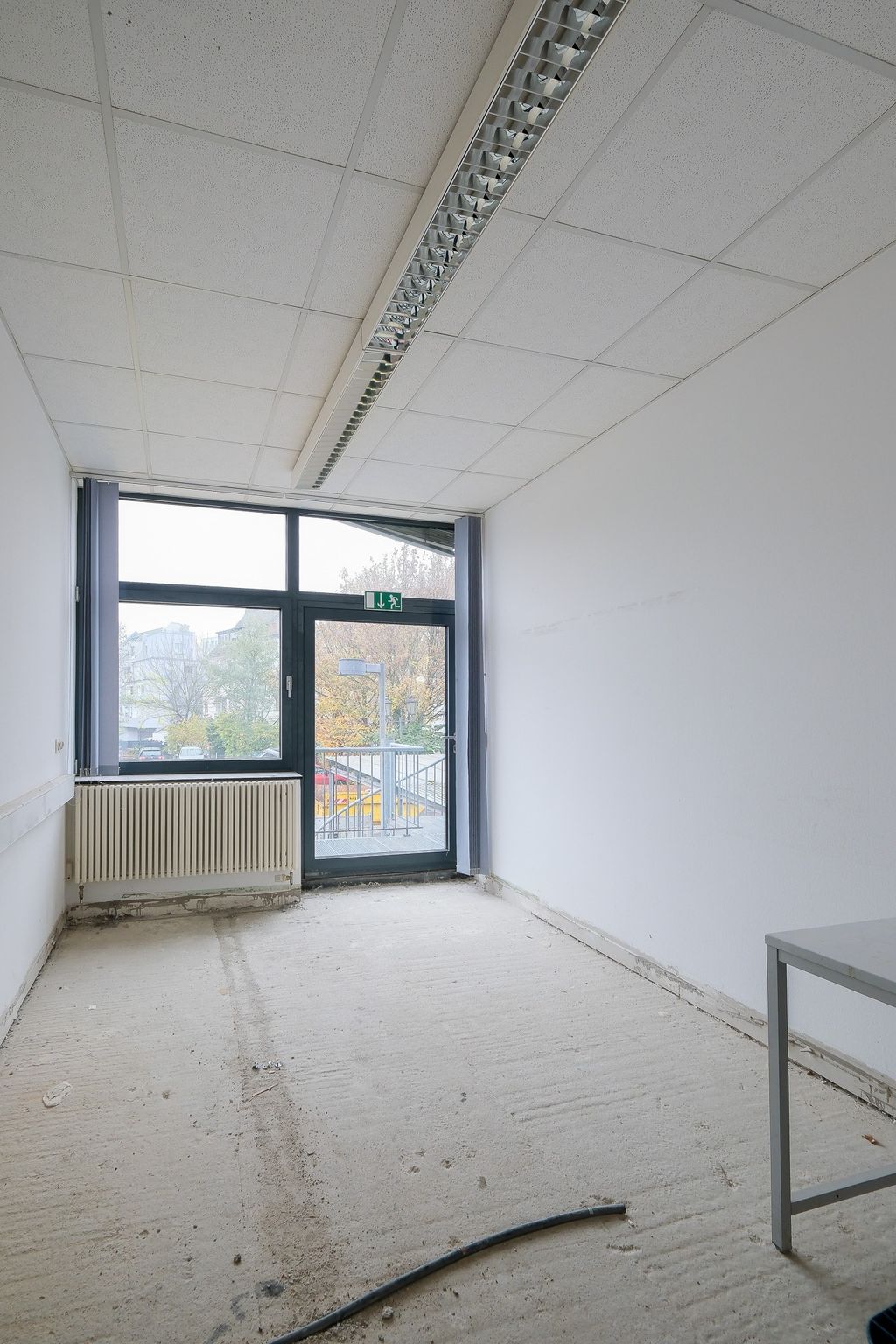 Leerer, unfertiger Raum mit nacktem Betonboden, weißen Wänden, einem großen Fenster und einer Glastür mit grünem Notausgangsschild, durch das man eine Außentreppe und Bäume sehen kann.