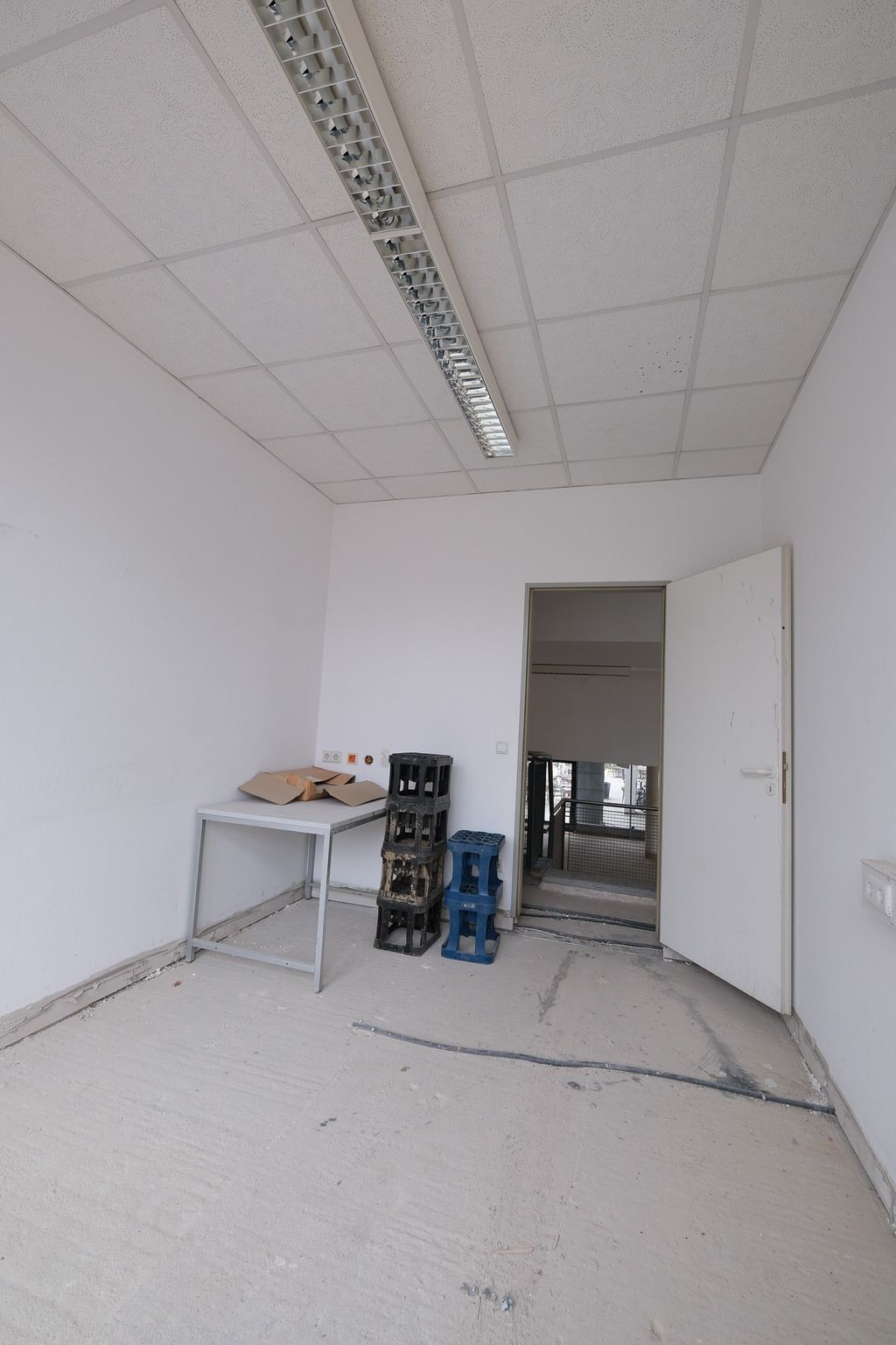 Leerer, unfertiger Raum mit einem weißen Tisch, drei gestapelten Plastikboxen und einer offenen Tür, die zu einem Treppenhaus führt.