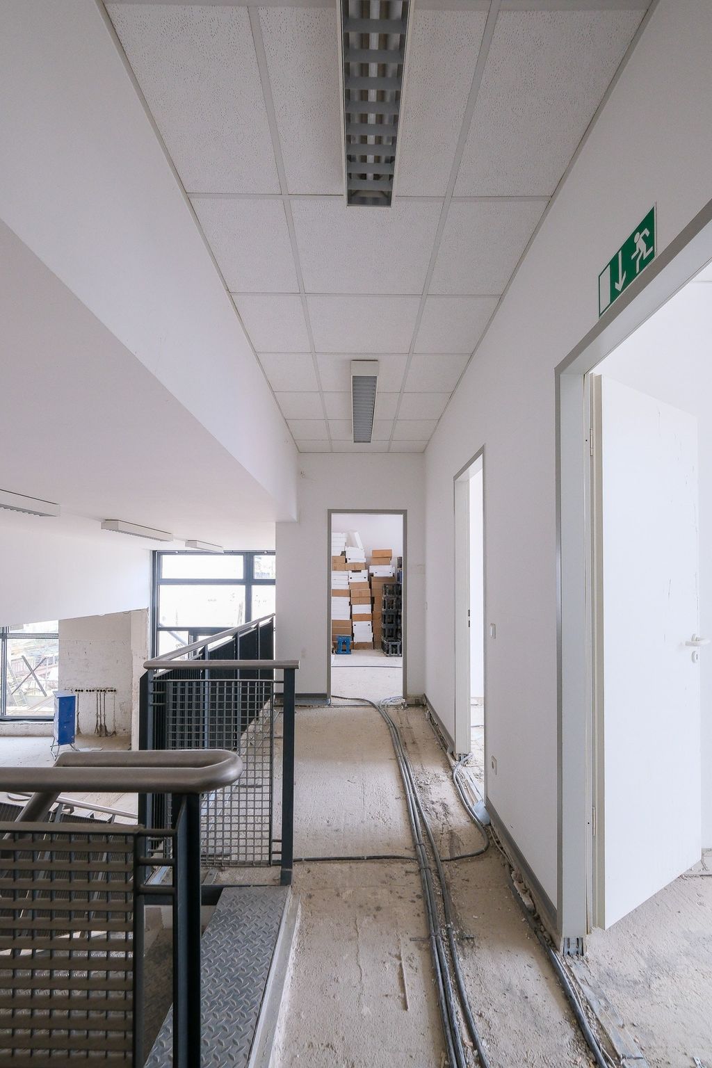 Innenansicht eines roh ausgebauten Flurs mit offenem Treppengeländer, sichtbaren Kabeln am Boden und mehreren Türen, von denen eine zu einem Raum mit gestapelten Kartons führt.