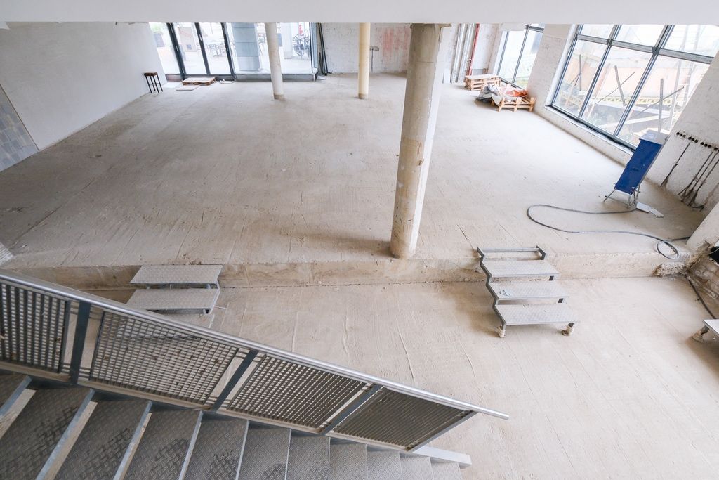 Innenraum einer unfertigen Baustelle mit rohem Boden, Treppen und mehreren Säulen, großes Fenster im Hintergrund mit Blick auf Außengerüst.