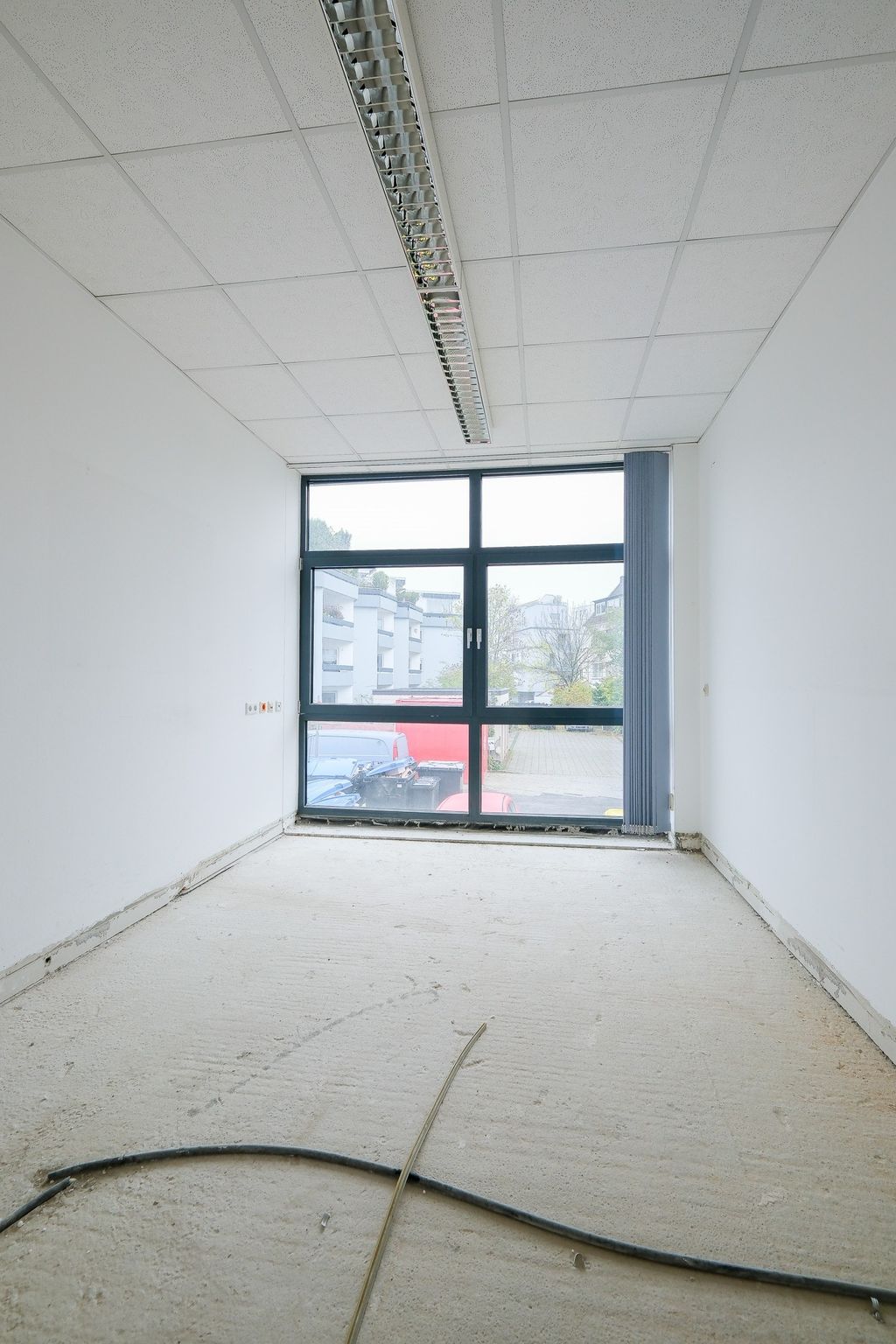 Leerer, unrenovierter Raum mit weißen Wänden und einem großen Fenster mit Blick auf außen parkende Fahrzeuge und Gebäude.