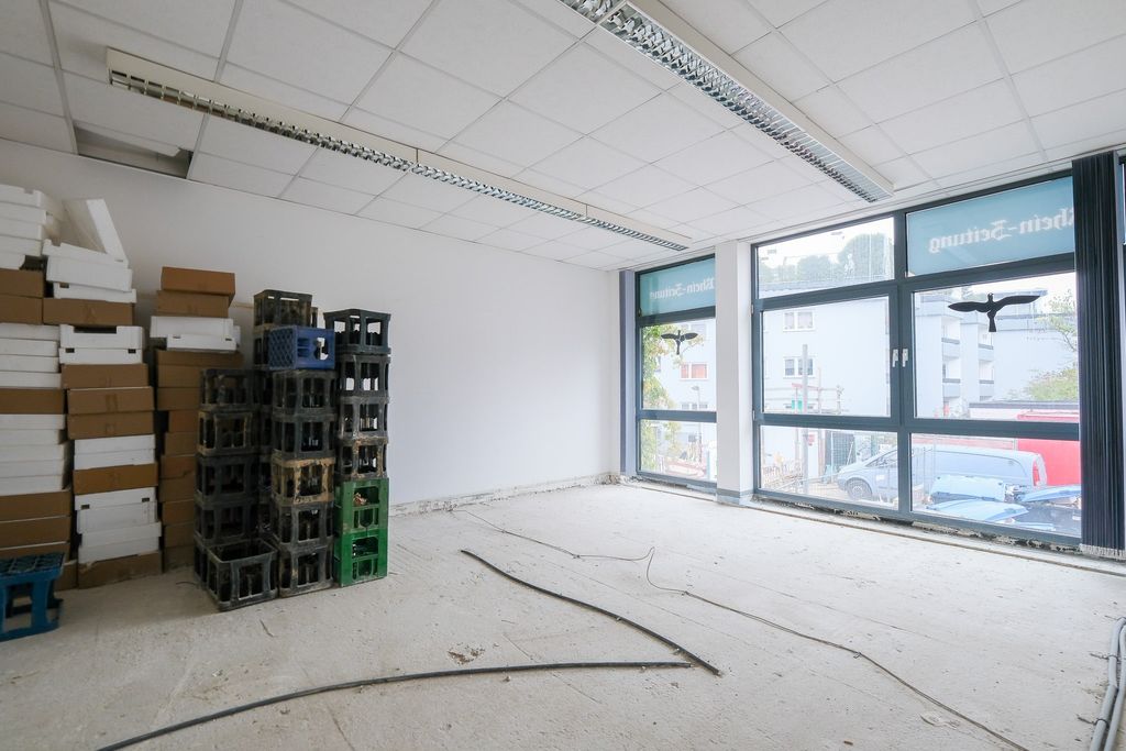 Leeres, renovierungsbedürftiges Zimmer mit großen Fenstern und gestapelten Pappkartons und Getränkekisten an einer Wand.