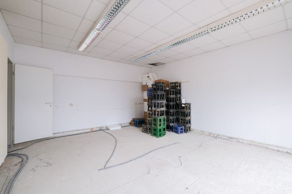 Leerer Raum mit weißen Wänden und Decke, an der Ecke sind mehrere gestapelte Kunststoffkisten und einige Kartons, offenes Türblatt an der linken Wand.