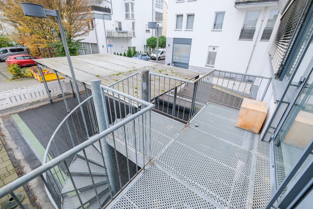 Hoch gelegene rechteckige Metallterrasse mit gelochtem Bodenbelag und umlaufendem Geländer, ein Holzblock steht in der Ecke, eine Wendeltreppe führt nach unten, umgeben von Wohngebäuden und Parkplätzen mit Herbstbäumen im Hintergrund.