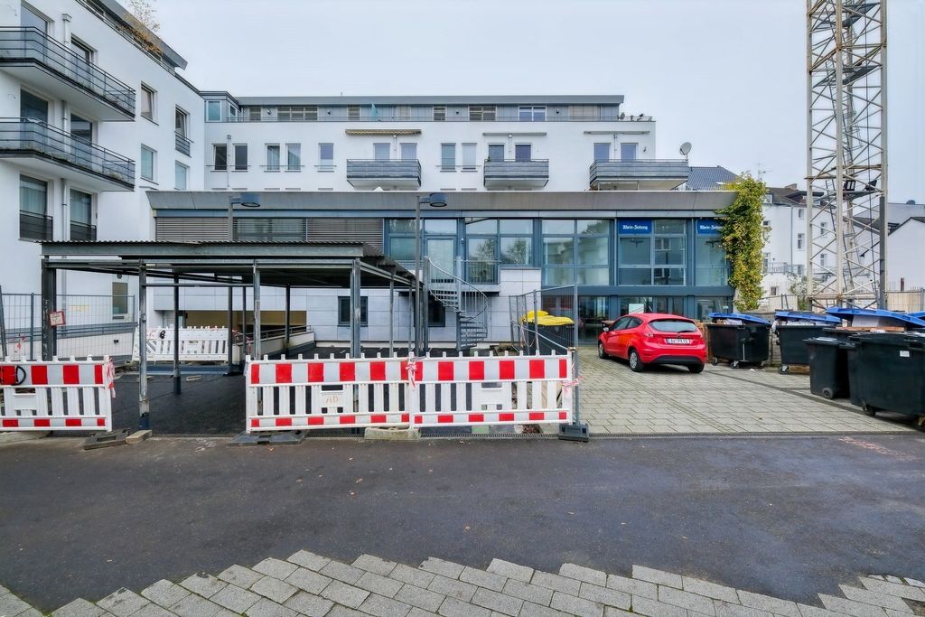 Moderne Wohngebäude mit hellen Fassaden und Balkonen im Hintergrund, davor eine Baustelle mit Absperrgittern und einer überdachten Fläche, rechts ein Parkplatz mit Müllcontainern und einem roten Auto.