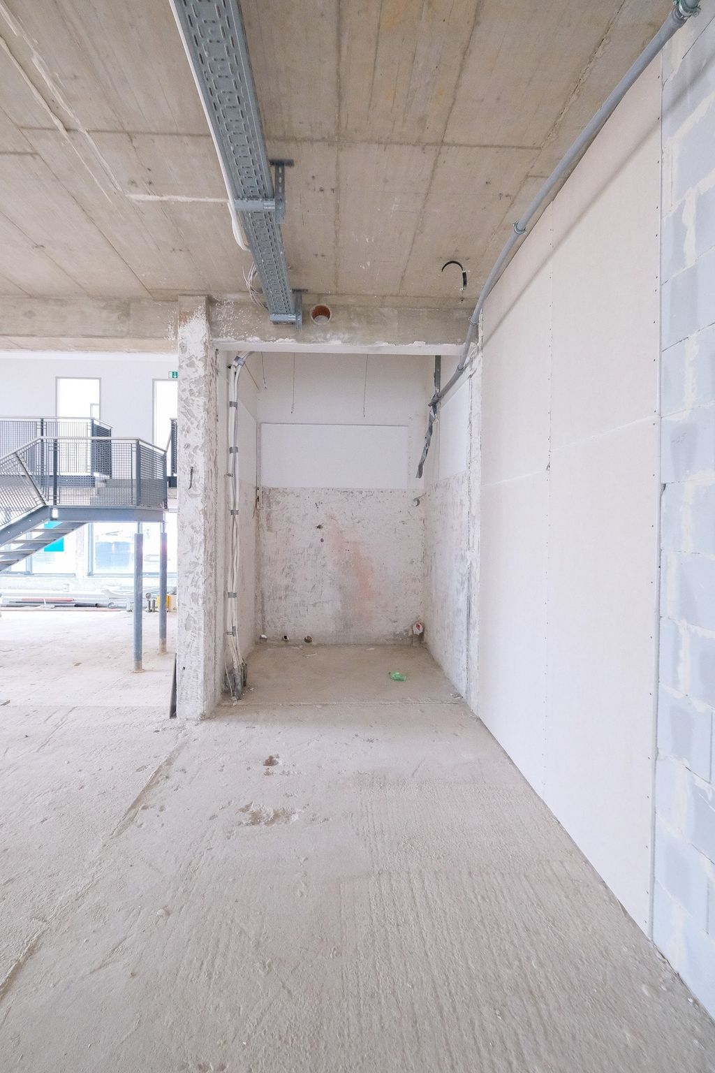 Rohbau eines Innenraums mit sichtbaren Betonböden, Wänden und Decke, teilweise mit Kabelkanälen und Treppe im Hintergrund.