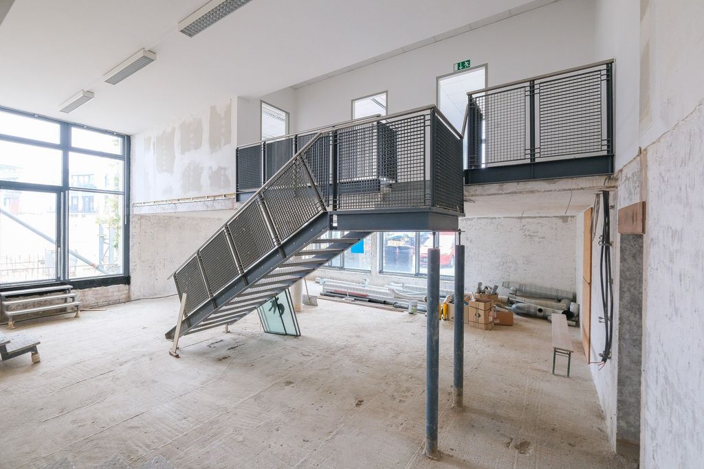 Innenraum eines leeren, unfertigen Gebäudes mit einer Metalltreppe, die zu einer Galerie führt. Große Fenster lassen Tageslicht herein, der Boden ist roh und Wände sind teilweise verputzt.