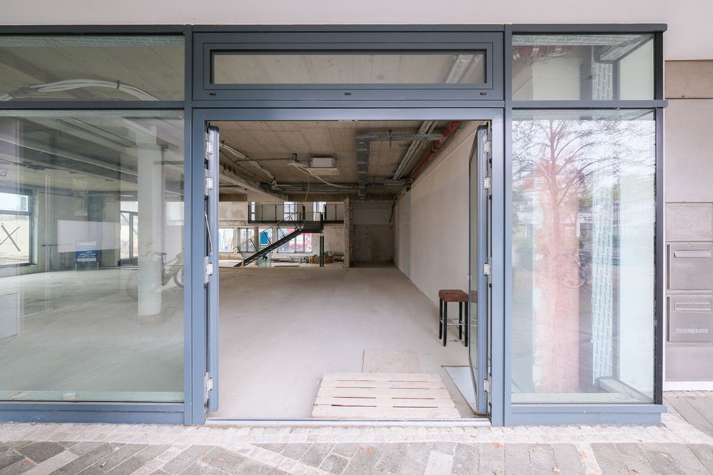 Leeres, modernes Gewerbeobjekt mit großen Fensterfronten und blauer Türrahmung, sichtbaren Rohren an der Decke und einer kleinen Holzpalette sowie einem Hocker im Inneren.