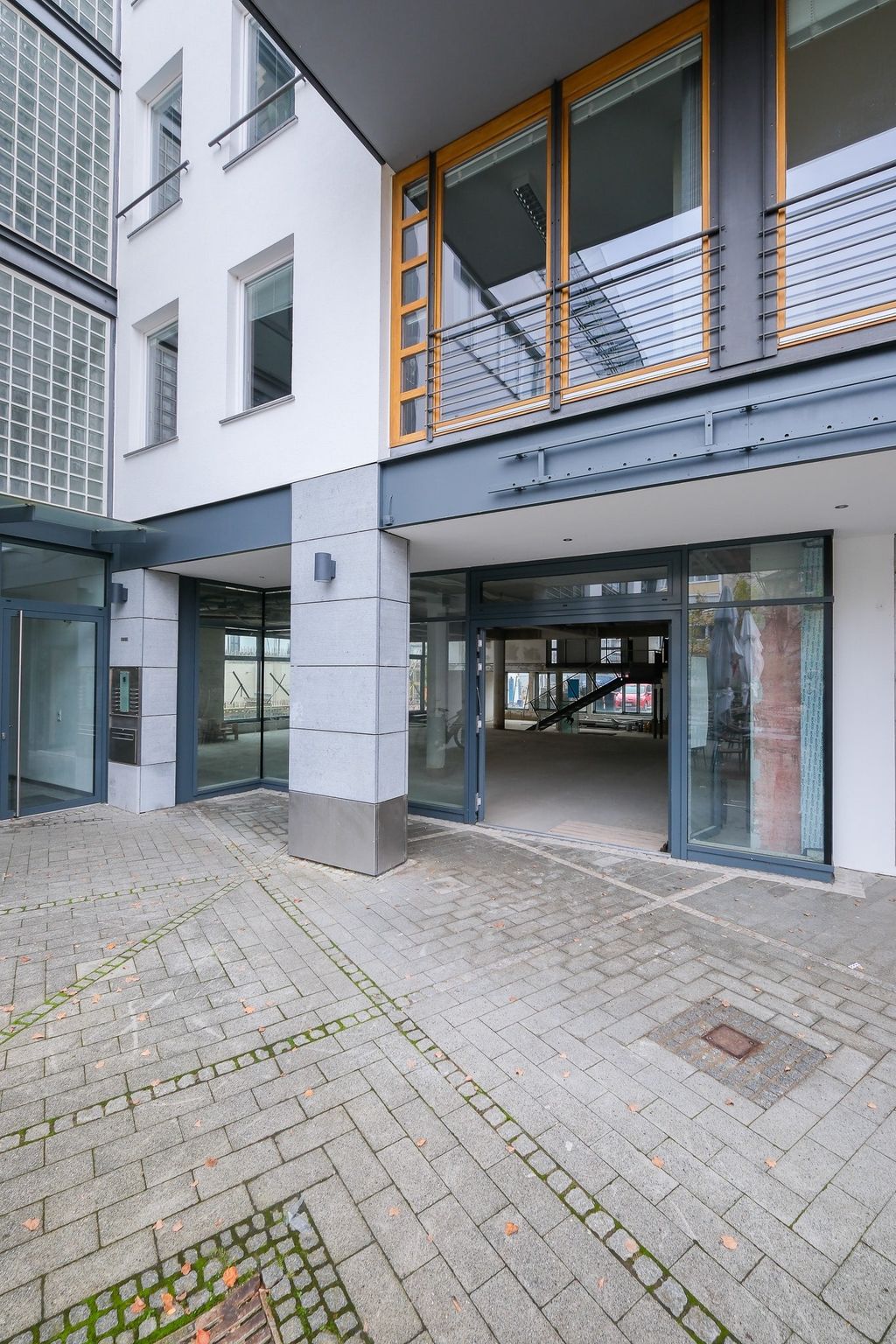 Eingangsbereich eines modernen Gebäudes mit großen Glasflächen und grauen Pflastersteinen.