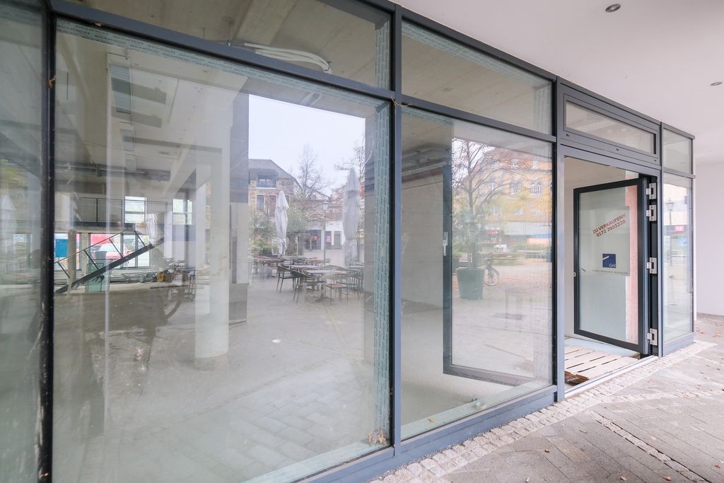 Spiegelfensterausschnitt eines modernen Gebäudes mit Blick auf einen leeren Innenhof mit Tischen, Stühlen und geschlossenen Sonnenschirmen an einem bewölkten Tag.