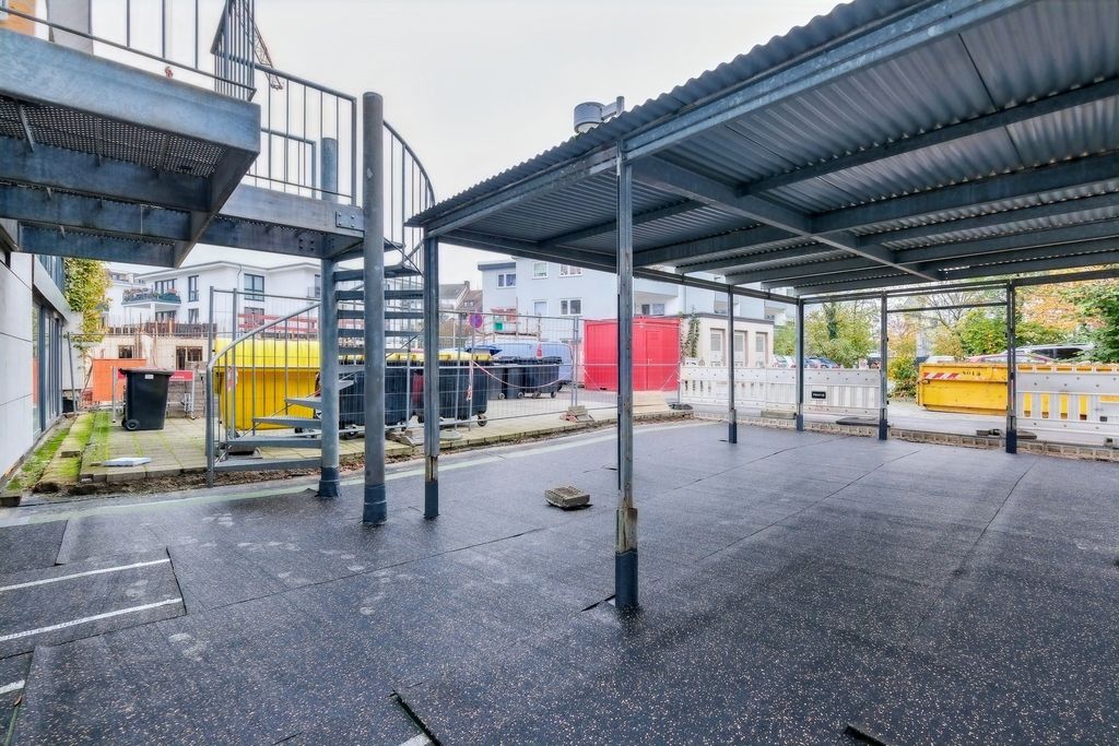 Überdachter Außenbereich mit einer Stahlkonstruktion und Spiraltreppe in einem urbanen Umfeld mit Müllcontainern und Baustellenelementen im Hintergrund.