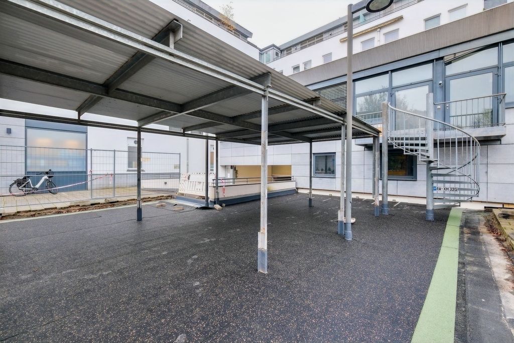 Außenbereich mit einem großen überdachten Bereich mit Metallstützen, einem vor Gebäuden platzierten Fahrrad und einer spiralförmigen Außentreppe an der Gebäudewand.