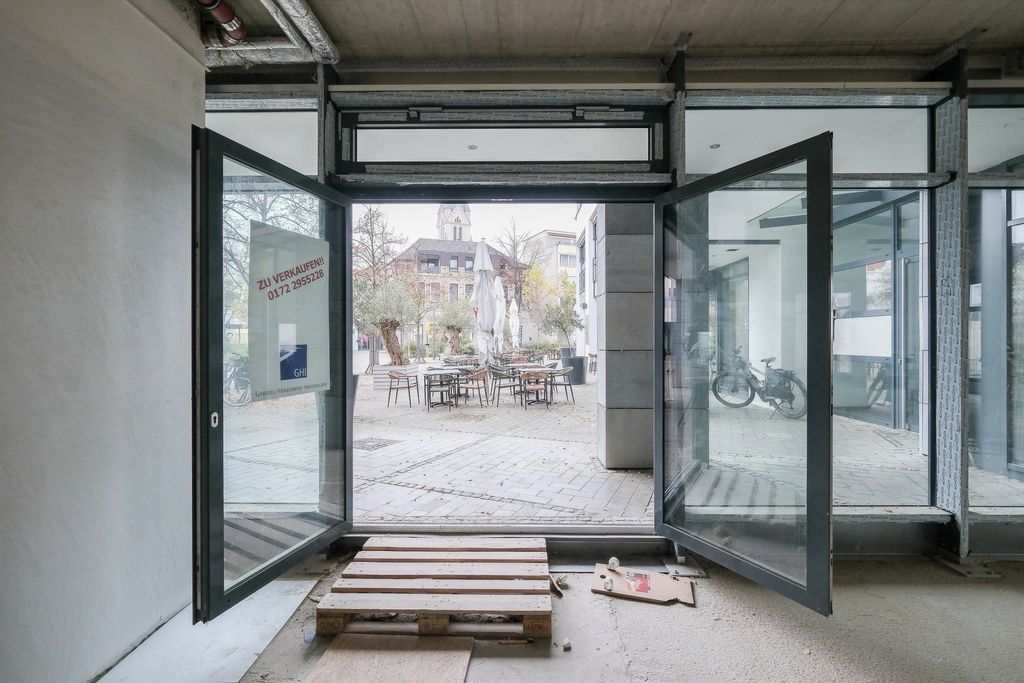 Blick durch eine offene Glastür auf einen gepflasterten Innenhof mit Stühlen, Tischen und einem geschlossenen Sonnenschirm, im Innenraum eine Holzpalette und eine Zettelbox auf dem Boden.