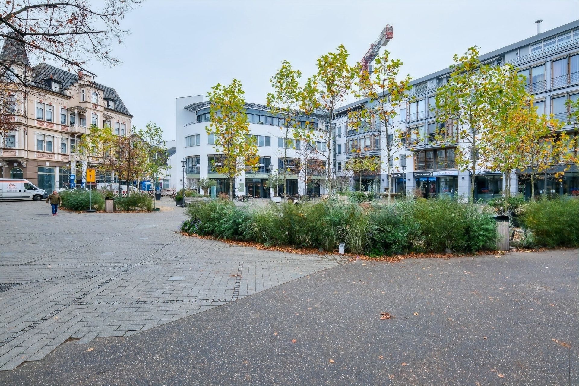 Urbaner Platz mit Bäumen, Sträuchern und modernen sowie älteren Gebäuden im Hintergrund.