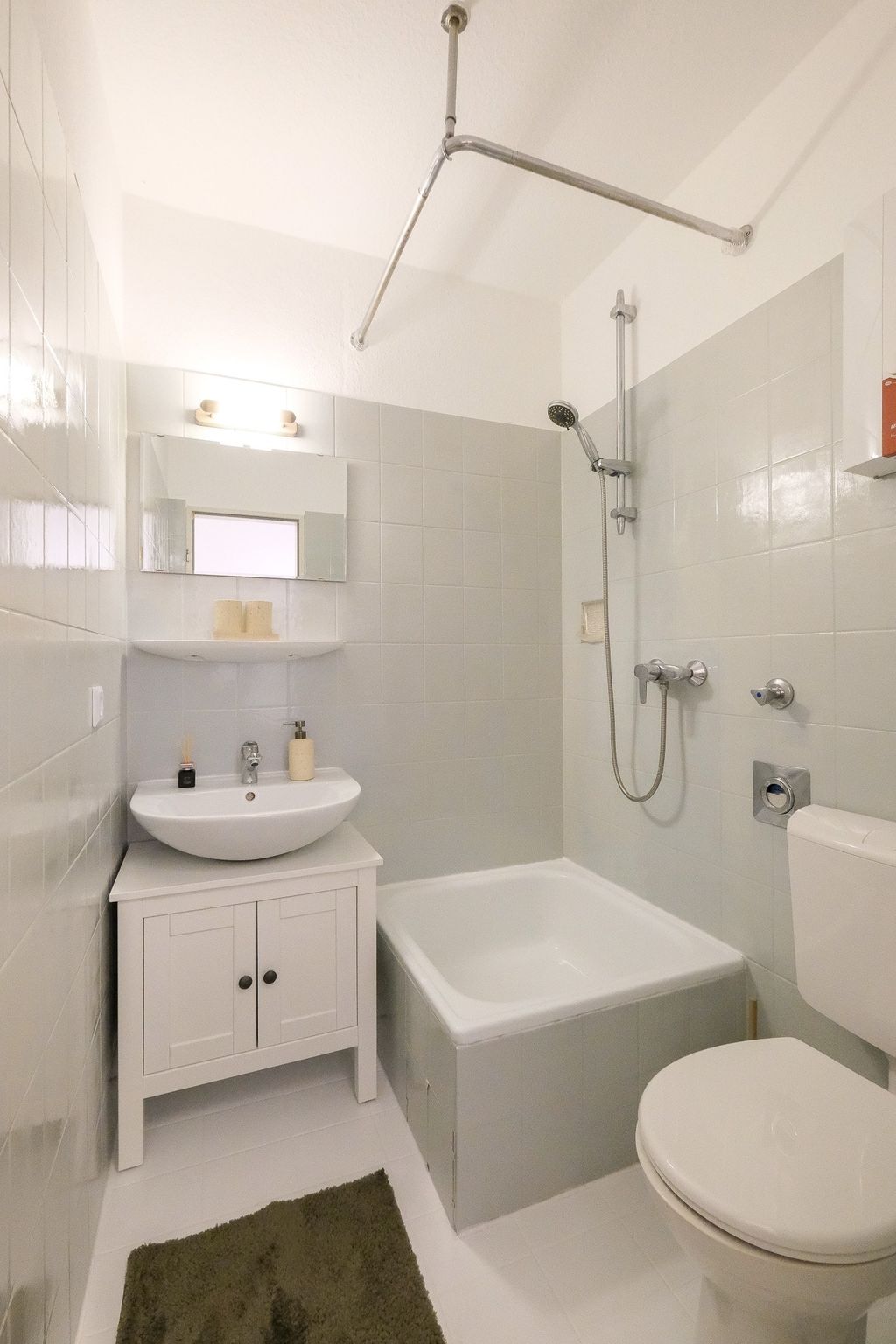 Kleines Badezimmer mit Waschbecken auf einem weißen Unterschrank, Toilette und Badewanne mit Duschkopf, grauen Fliesen und einem dunkelgrünen Badvorleger auf dem Boden.
