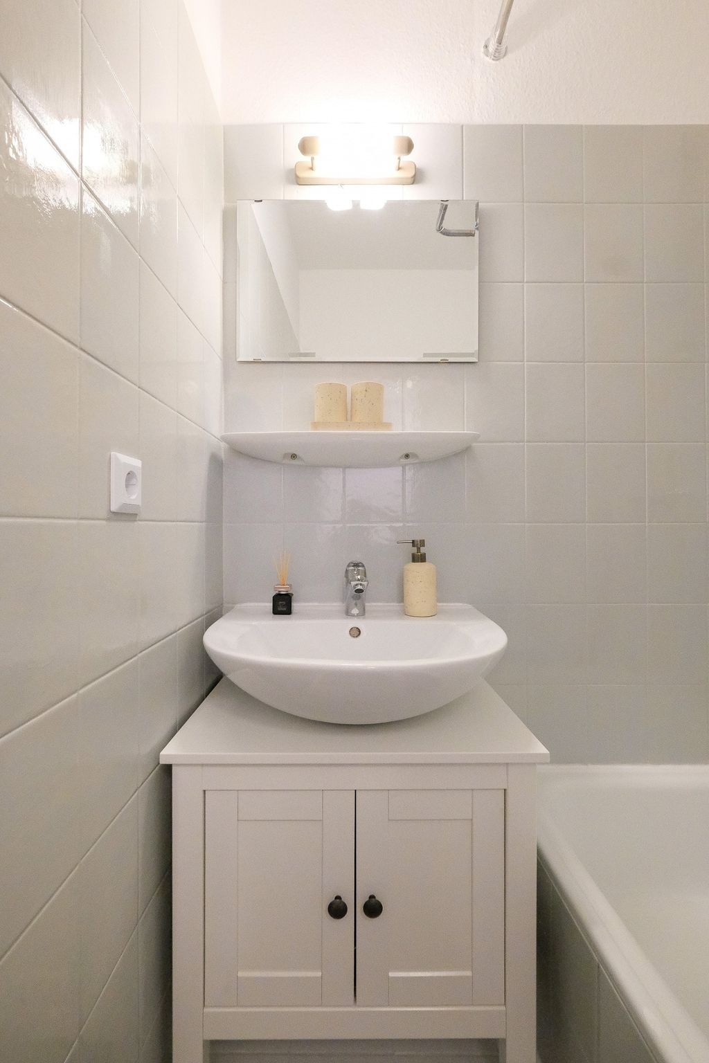 Kleines Badezimmer mit weißem Waschbecken auf einem weißen Schrank, darüber ein Spiegel und zwei Kerzen auf einem schwebenden Regal, umgeben von beige-weißen Fliesen.
