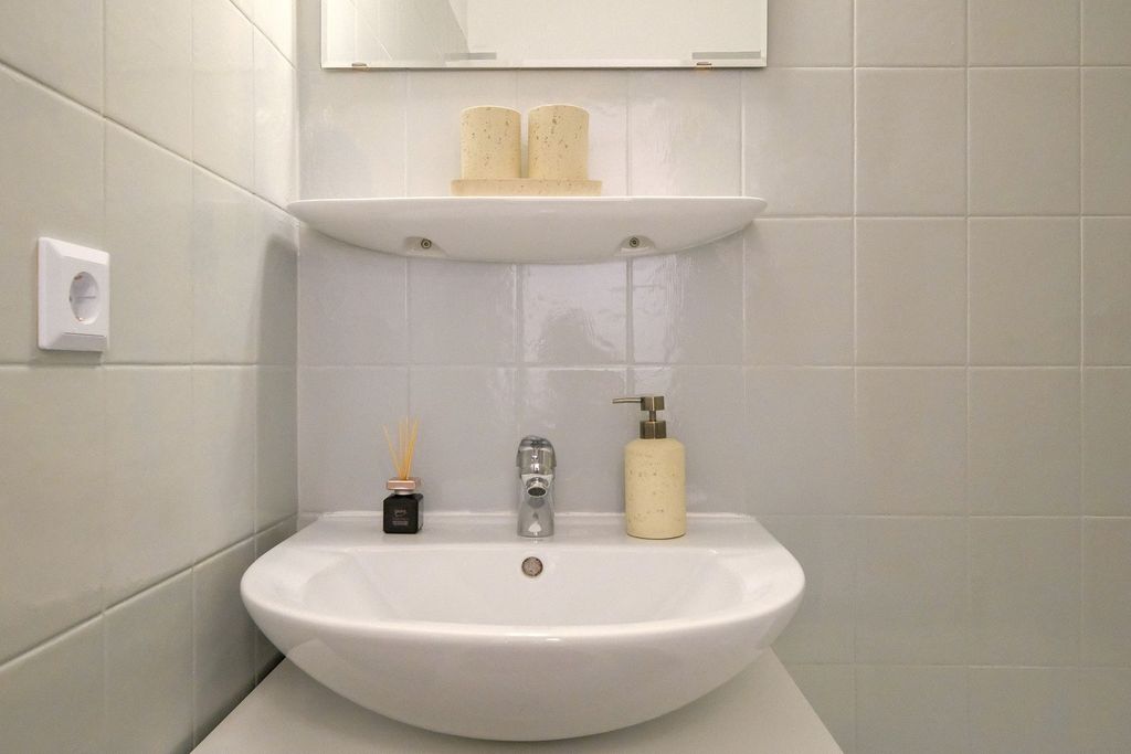 Weißes Waschbecken mit Chromhahn in einem Badezimmer mit hellgrauen Fliesen an der Wand, darauf stehen ein Seifenspender, ein Duftdiffusor und oben zwei Kerzen auf einem Regal unter einem Spiegel.