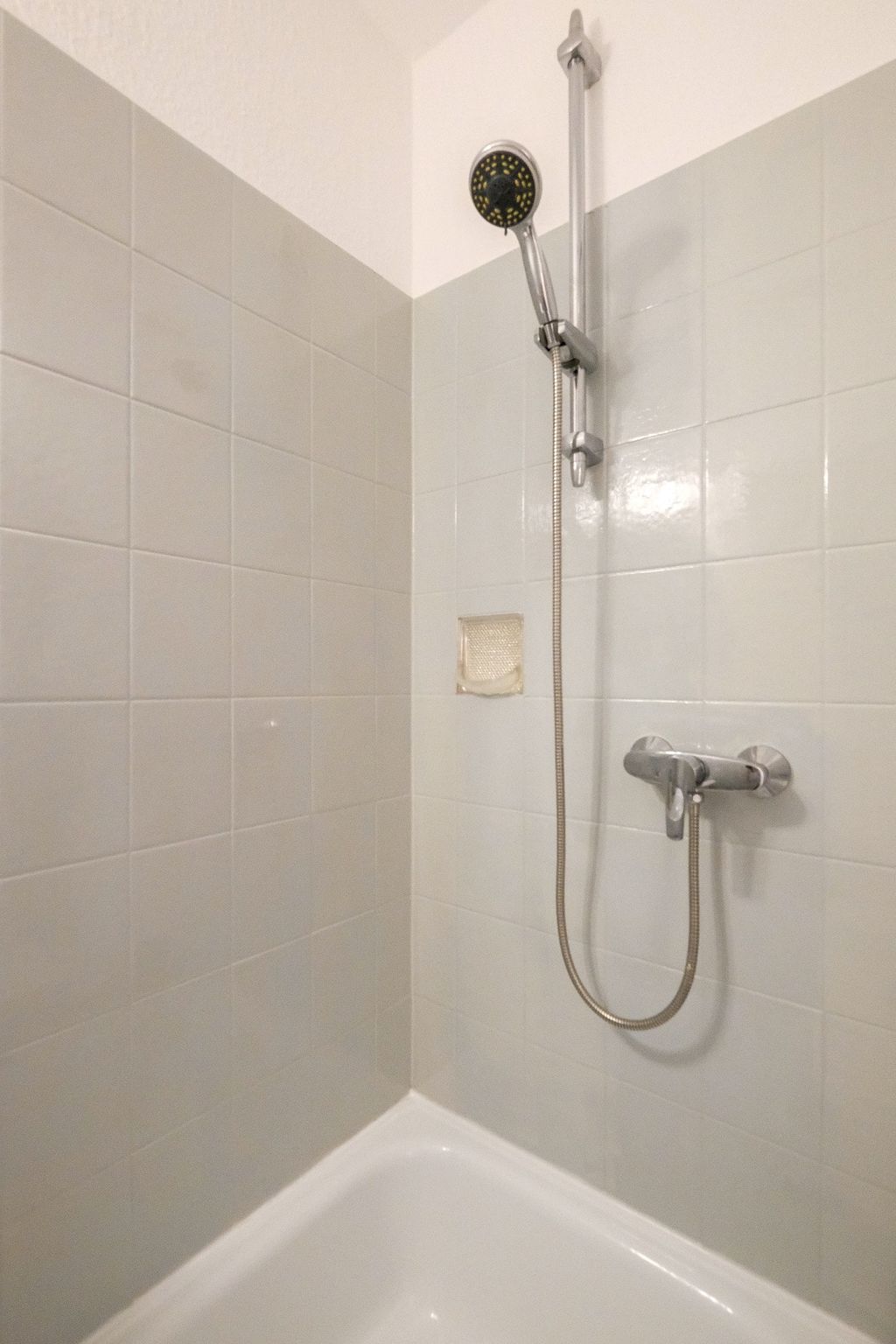 Ein einfache Duschkabine mit weißen Wandfliesen, einer verstellbaren Handbrause und einem Wasserhahn, über einer Badewanne.