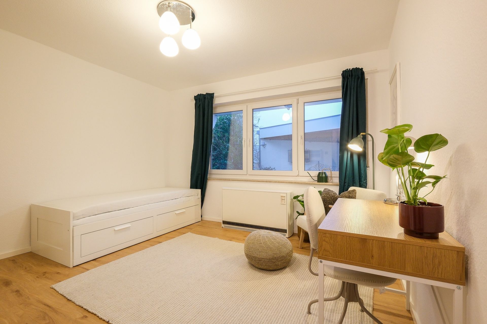 Hell eingerichtetes Zimmer mit einem Tagesbett mit Schubladen, einem Schreibtisch mit Pflanzen und einer sitzenden Stuhl sowie einem großen Fenster mit dunklen Vorhängen.