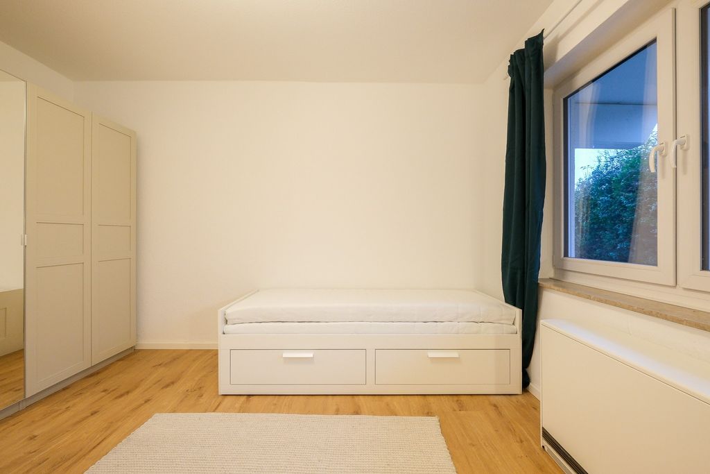Ein kleines, schlicht eingerichtetes Zimmer mit einem weißen Einzelbett mit zwei Schubladen, einem grauen Vorhang vor einem Fenster und einem großen Kleiderschrank mit Spiegel an der Wand.
