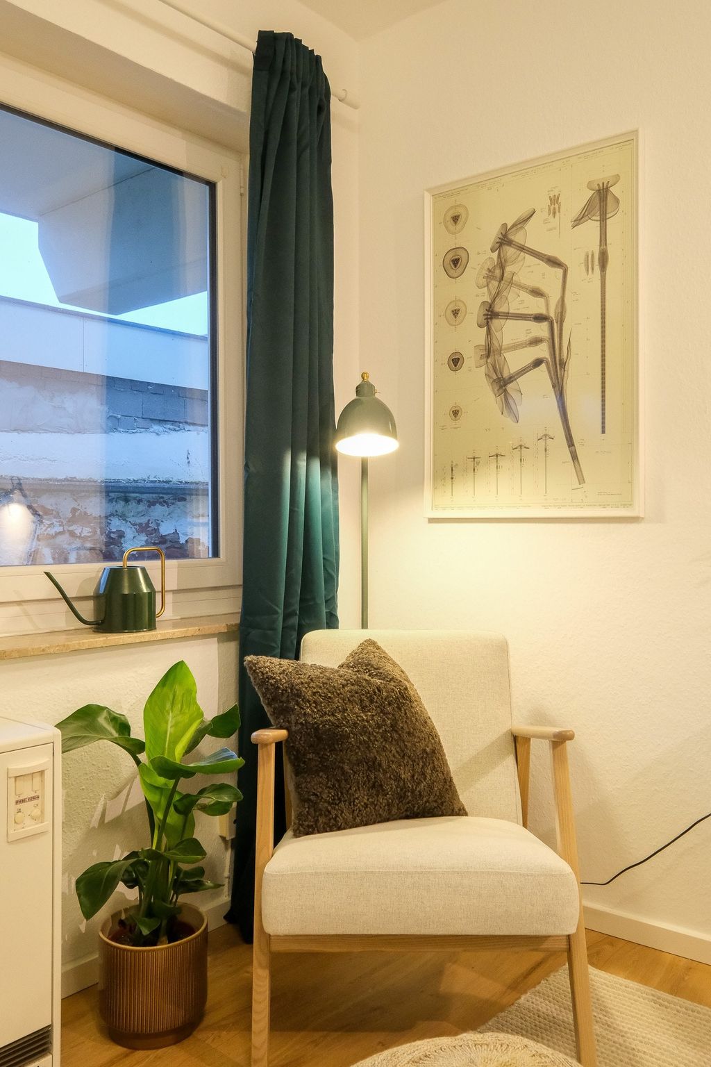 Ein gemütlicher Raum mit einem beigen Sessel, darauf ein braunes Kissen, neben einem Topfpflanze und einem Fenster mit blauen Vorhängen sowie einer grünen Stehlampe und einem botanischen Poster an der Wand.