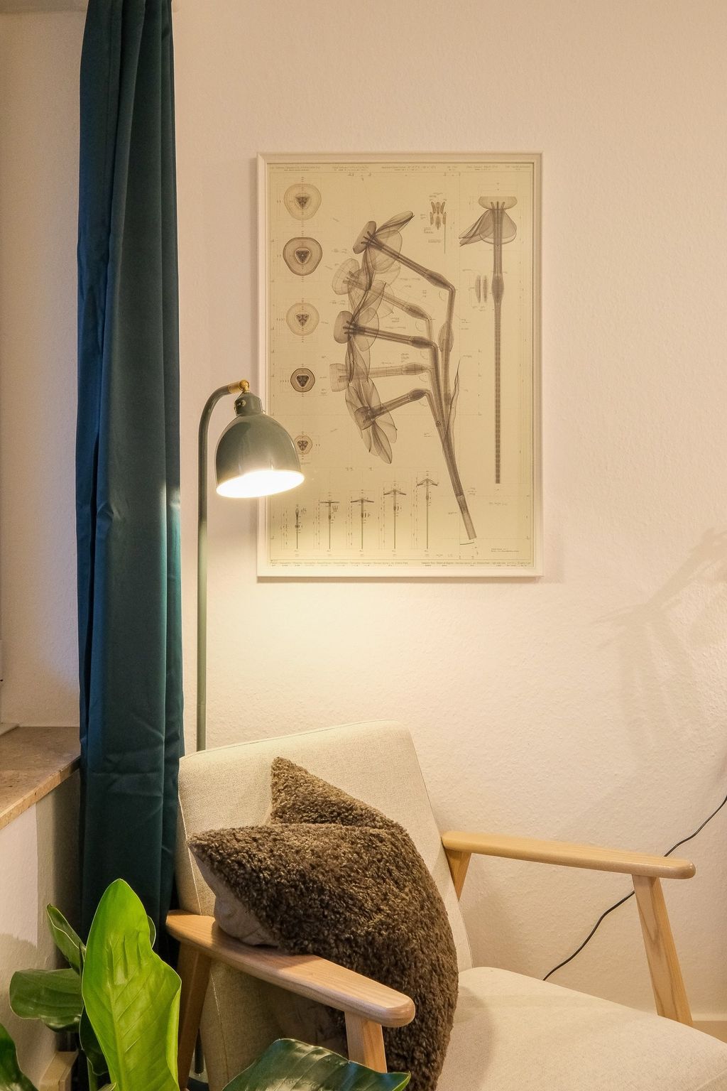 Ein gemütlicher Sessel mit zwei braunen Kissen neben einer grünen Stehlampe und einem großen botanischen Poster an der Wand.