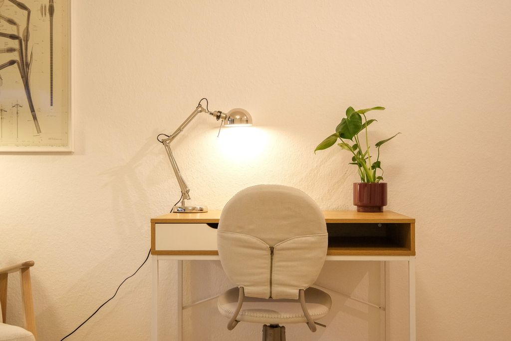 Ein einfacher Schreibtisch mit einer verstellbaren Schreibtischlampe, einem Topfpflanze und einem beigen Bürostuhl vor einer weißen Wand.