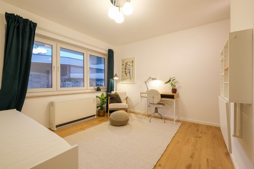Ein gemütliches Zimmer mit hellen Wänden, Parkettboden, einem hellen Schreibtisch mit Stuhl und Lampe, einem Sessel mit Kissen, einem Pflanzenkübel, großen Fenstern mit dunkelgrünen Vorhängen und einem hellen Teppich auf dem Boden.