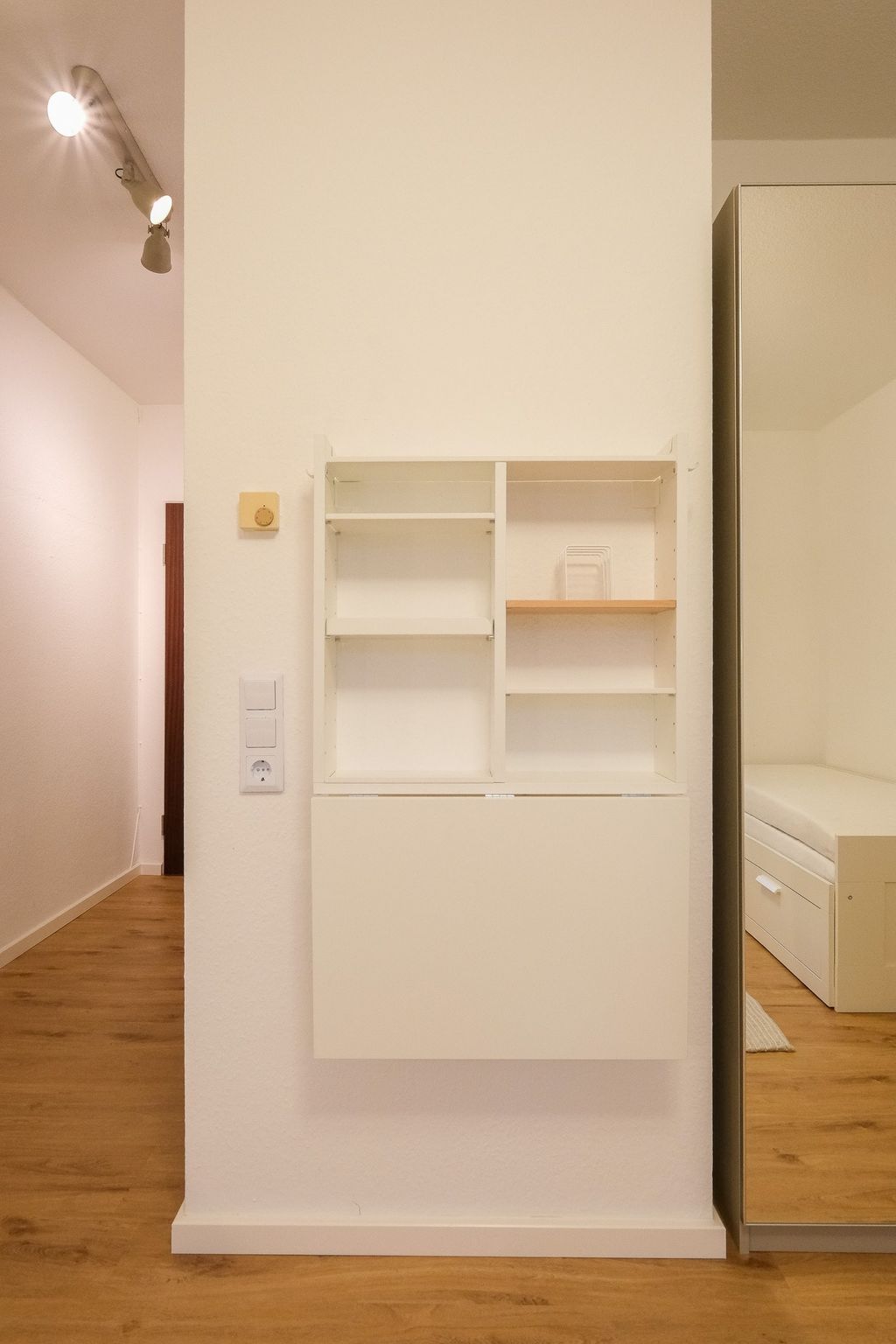 Weiße Wand mit einer kleinen weißen Regalbox und einem Spiegel daneben, Parkettboden und schlichte Deckenbeleuchtung im Flur eines modernen Raums.