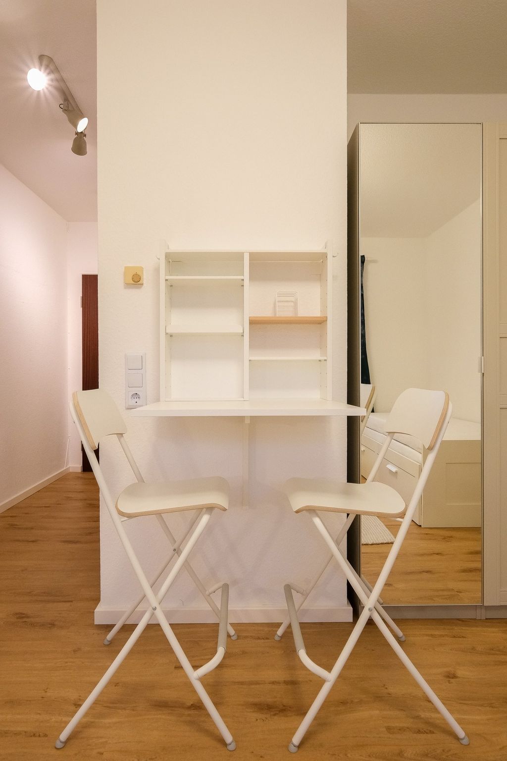 Kleiner Essbereich mit weißen Klappstühlen und einem an der Wand montierten weißen Klapptisch in einem modernen, hellen Raum mit Holzboden.