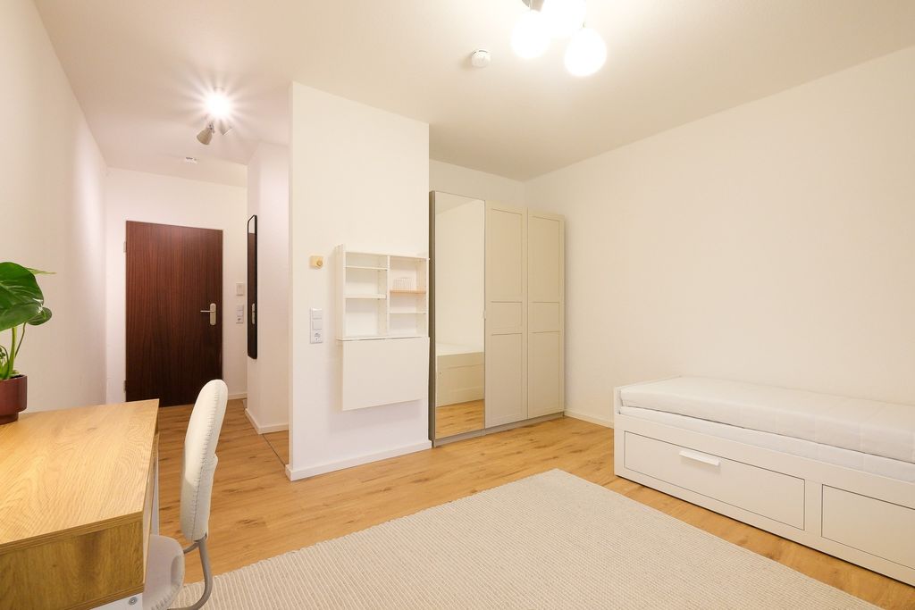 Helles, minimalistisch eingerichtetes Zimmer mit Holzboden, einem Schreibtisch mit Stuhl links und einem Bett mit Stauraum rechts, weiße Wände und ein großer Spiegel an einem Kleiderschrank.