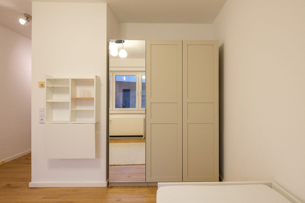 Ein Schlafzimmer mit einem weißen Doppelkleiderschrank, wobei eine der Türen verspiegelt ist. An der linken Wand sind offene Wandregale angebracht und im Vordergrund ist ein Teil eines Bettes sichtbar.