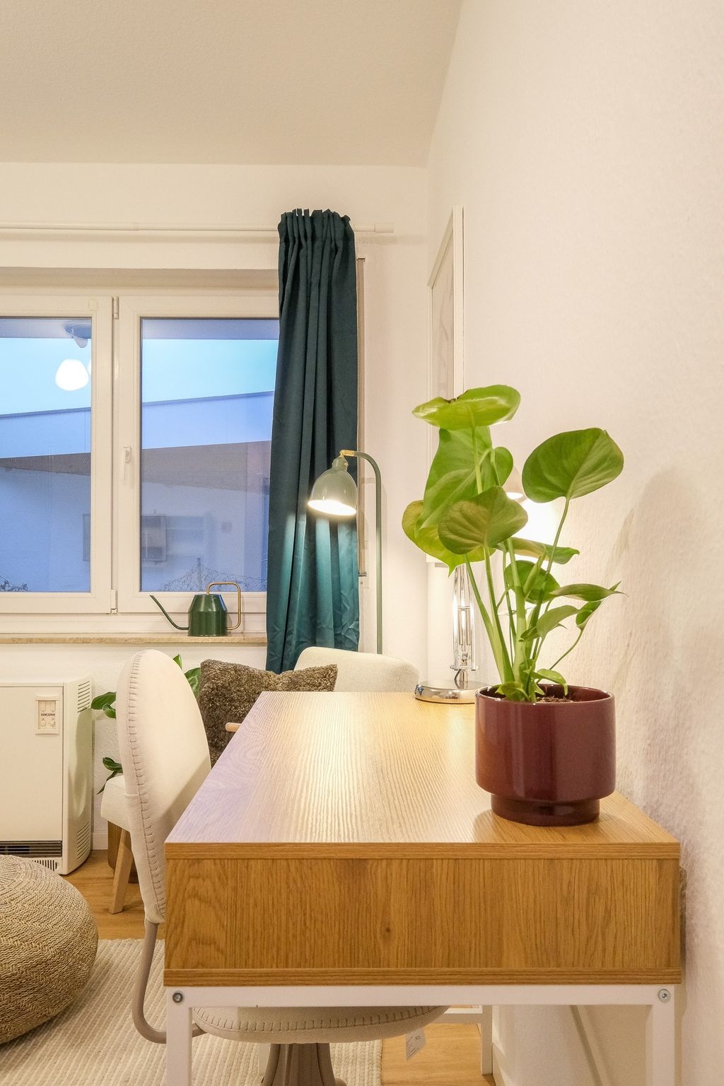 Moderner Schreibtisch aus hellem Holz vor einer weißen Wand mit einer Topfpflanze, einer Tischlampe und einem Fenster mit dunkelblauem Vorhang im Hintergrund.