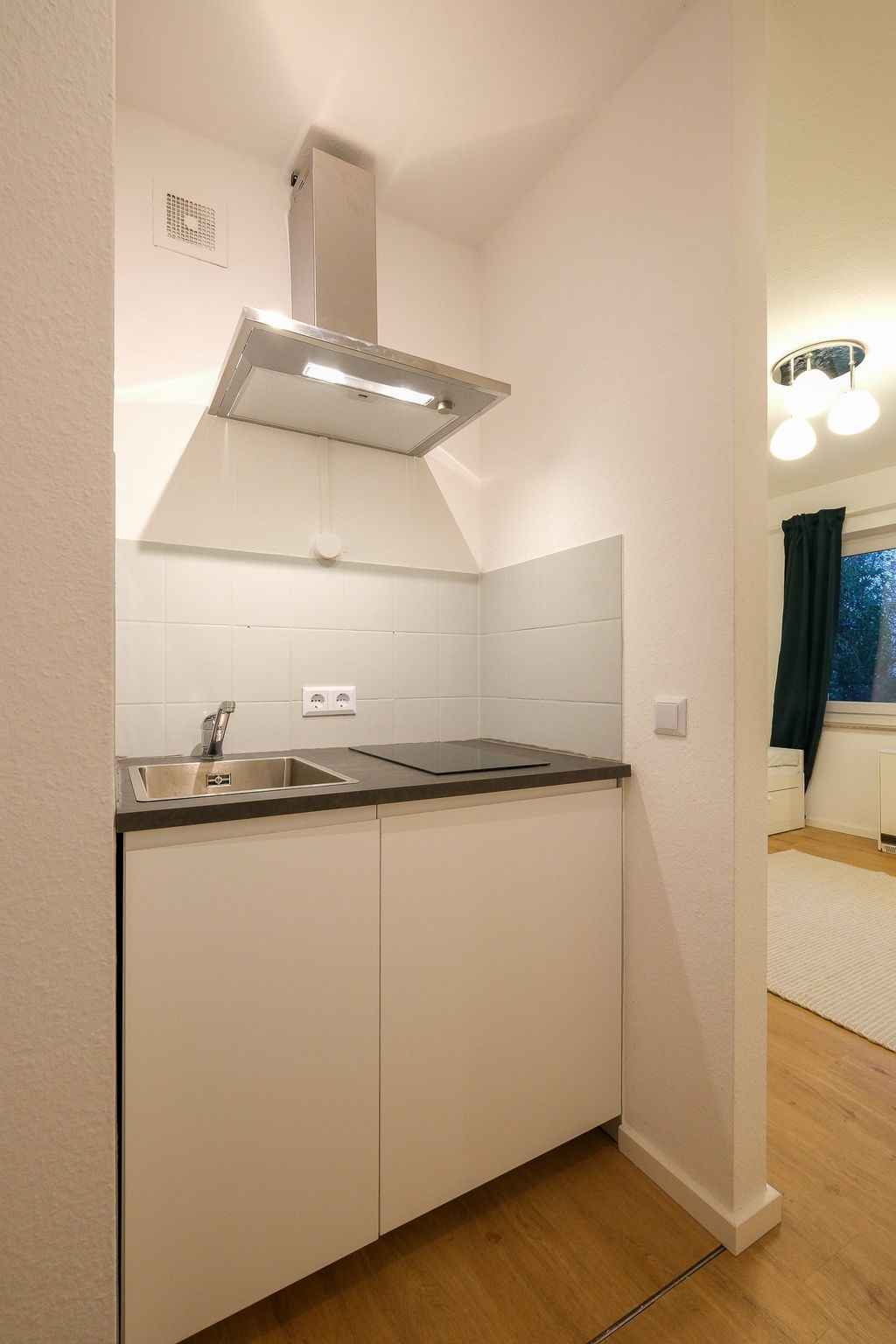 Kleine, moderne Küchenzeile mit Spüle, Induktionskochfeld und Edelstahl-Dunstabzug in einem weißen Raum mit Holzboden.