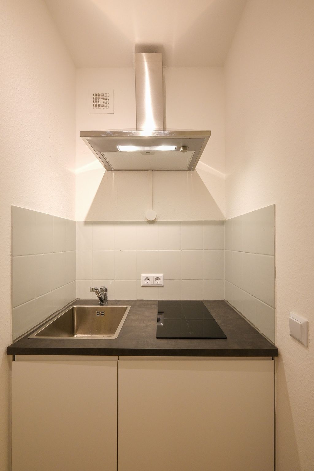 Kleine moderne Küche mit Spüle, Kochfeld und Edelstahl-Dunstabzug in einer Nische mit weißen Fliesen.