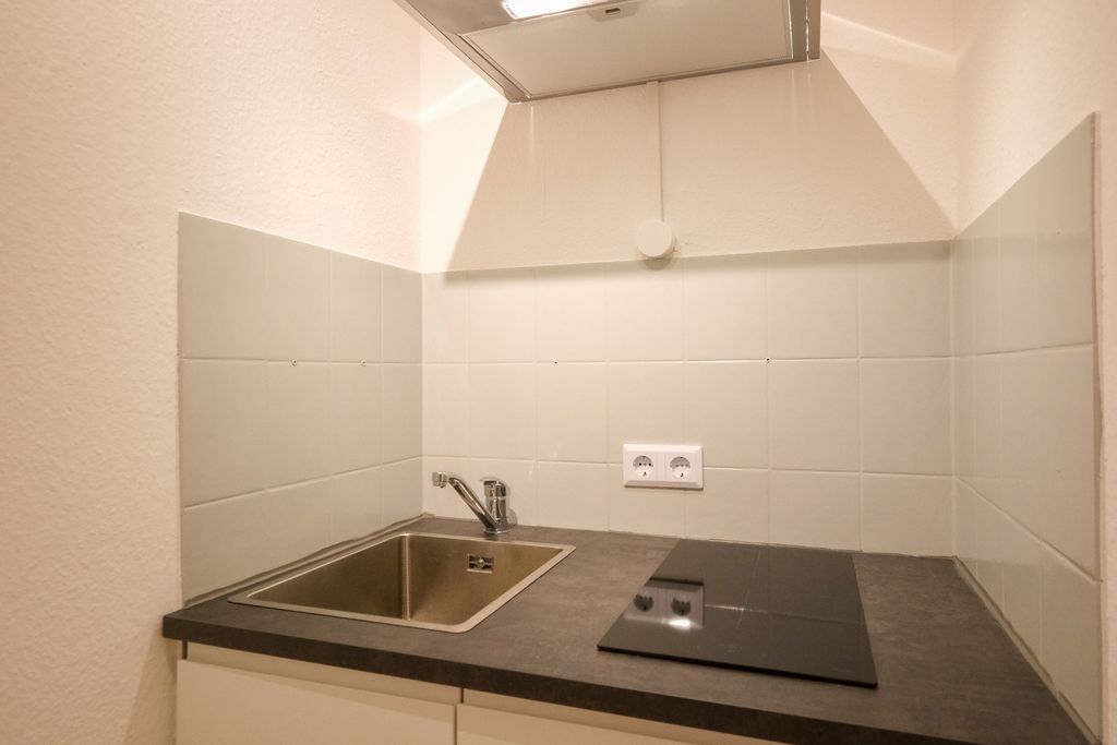 Kleine Küchenzeile mit Edelstahlspüle, Cerankochfeld und weiß gefliester Wand im Hintergrund.