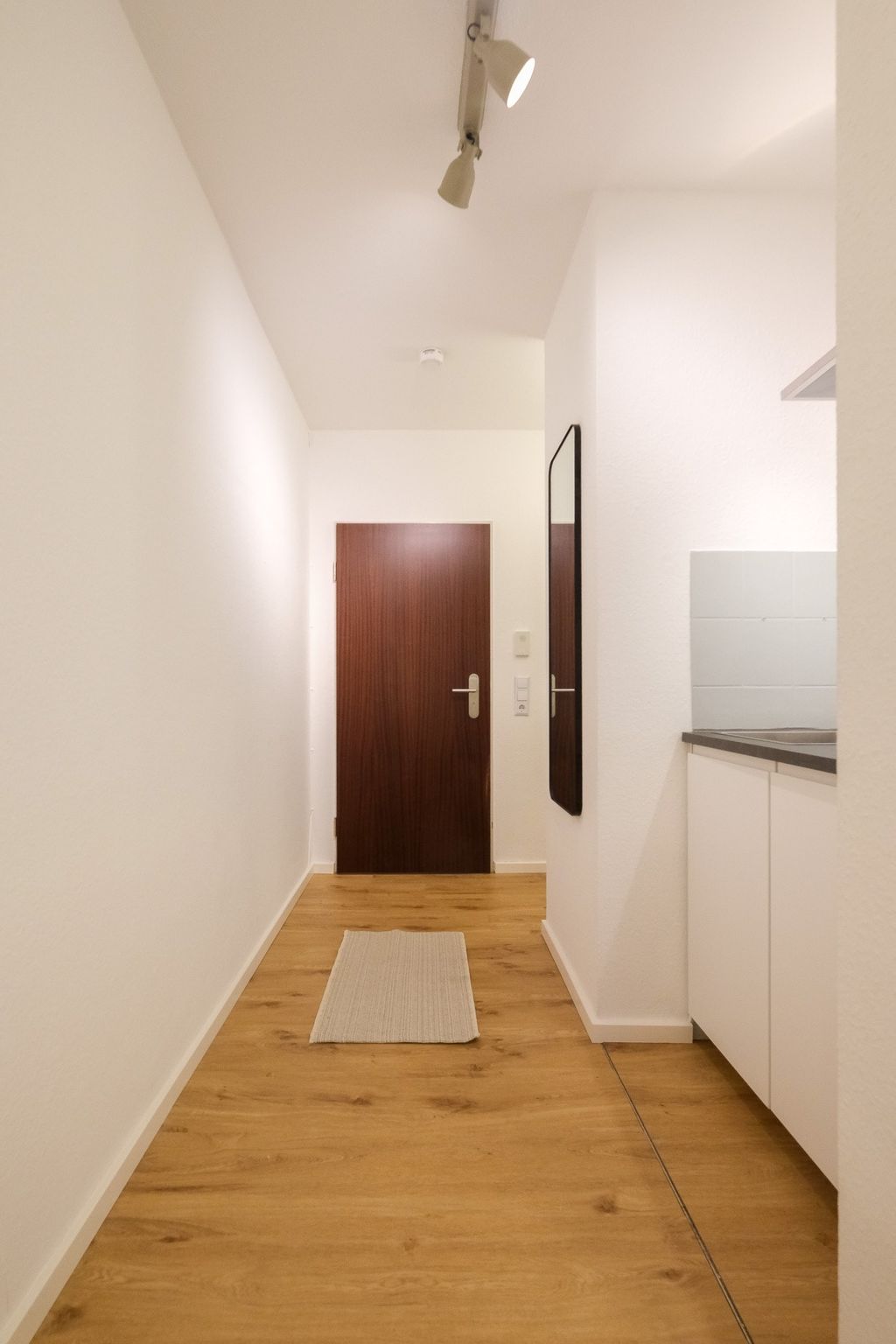 Schmaler Flur mit hellen Wänden, Holzboden und einem kleinen Teppich in der Mitte, am Ende eine dunkle Tür und seitlich ein rechteckiger Spiegel.