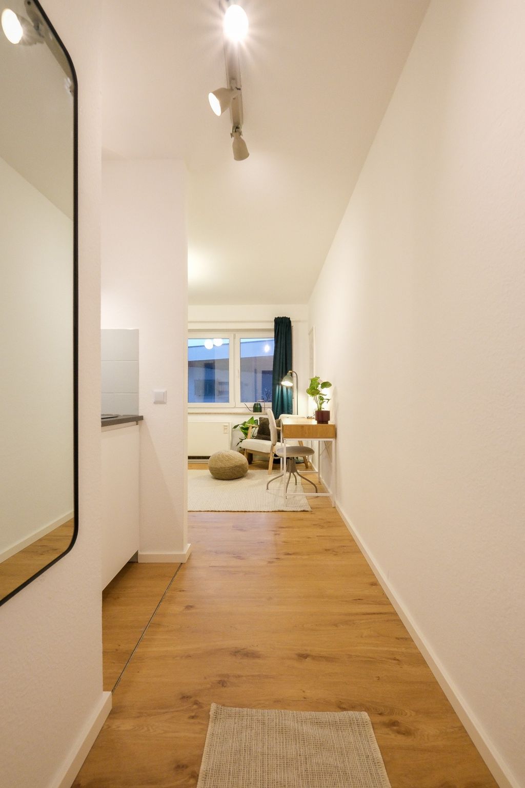 Ein heller Flur mit Holzboden führt zu einem gemütlichen Bereich mit Sesseln, einem Pouf, einem Schreibtisch mit einer Lampe und Pflanzen vor einem Fenster mit dunkelgrünen Vorhängen.