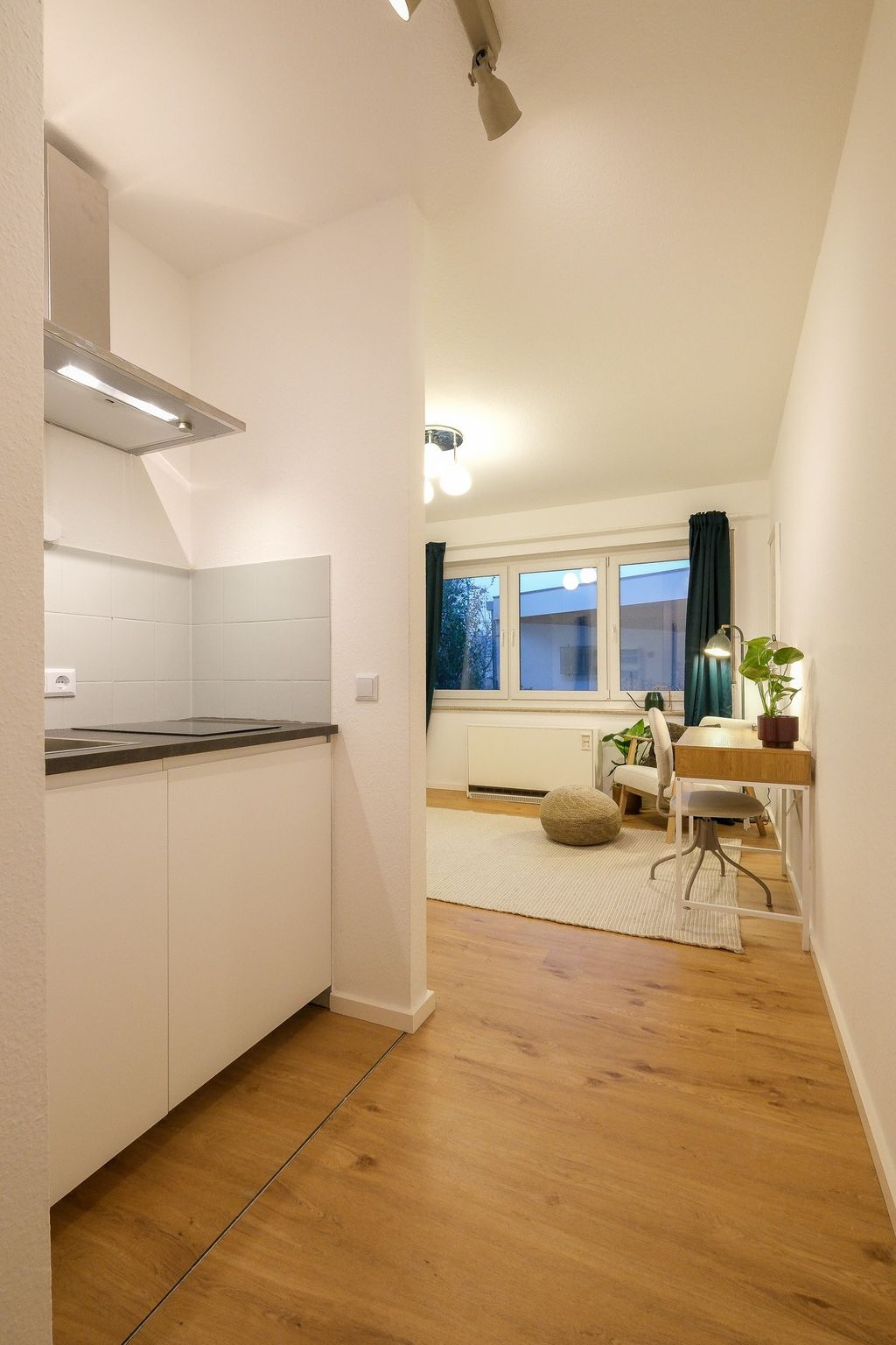 Ein heller Raum mit Laminatboden, einer kleinen weißen Küchenzeile links und einem Arbeitsbereich mit Schreibtisch, Stuhl, Pflanzen und Fenster mit blauen Vorhängen im Hintergrund.