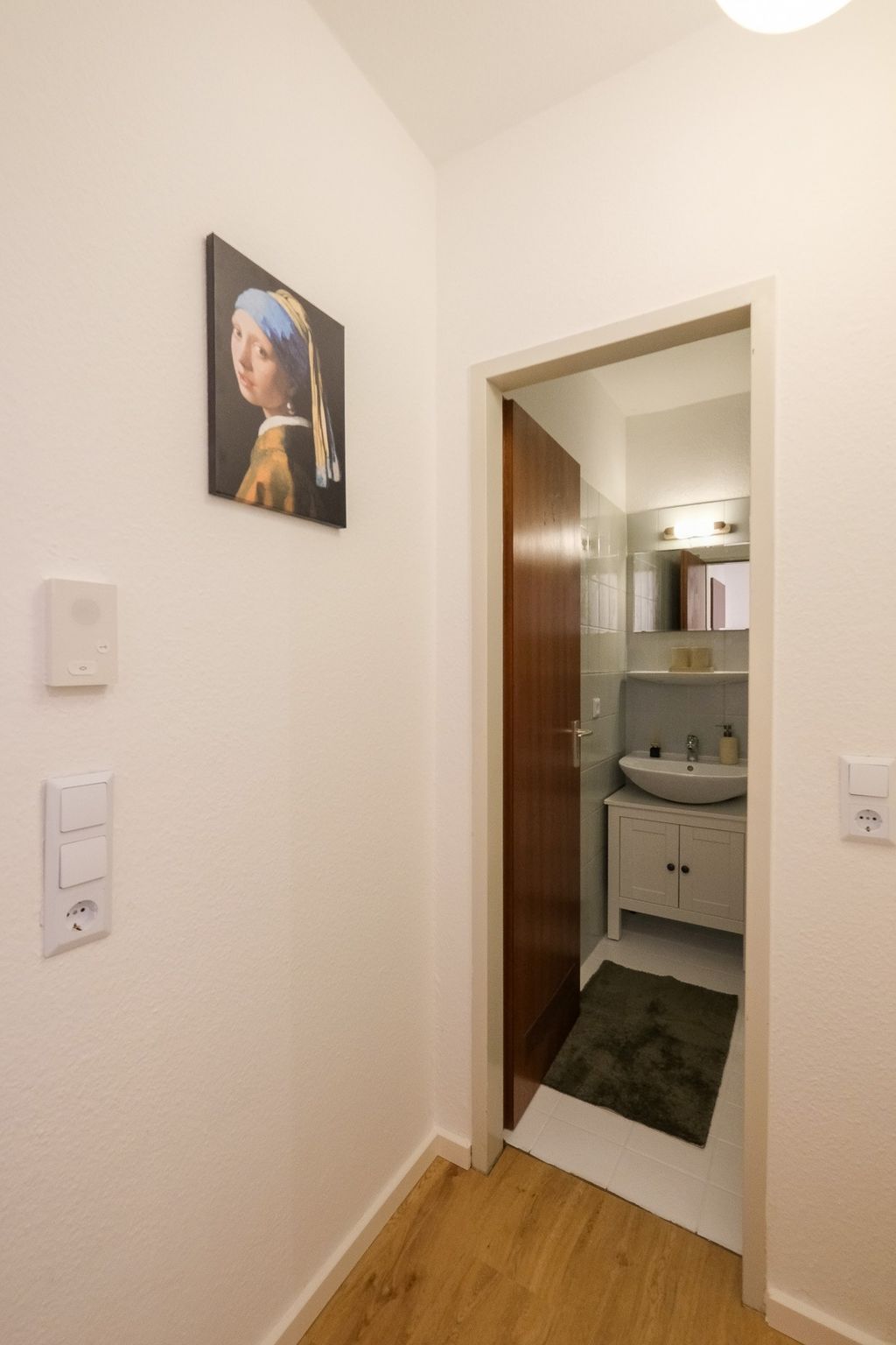 Blick in einen kleinen Waschraum mit geöffnetem Holztür, weißem Einrichtungsschrank und grauer Badematte, an der Wand rechts ein Gemälde des Mädchens mit dem Perlenohrring.