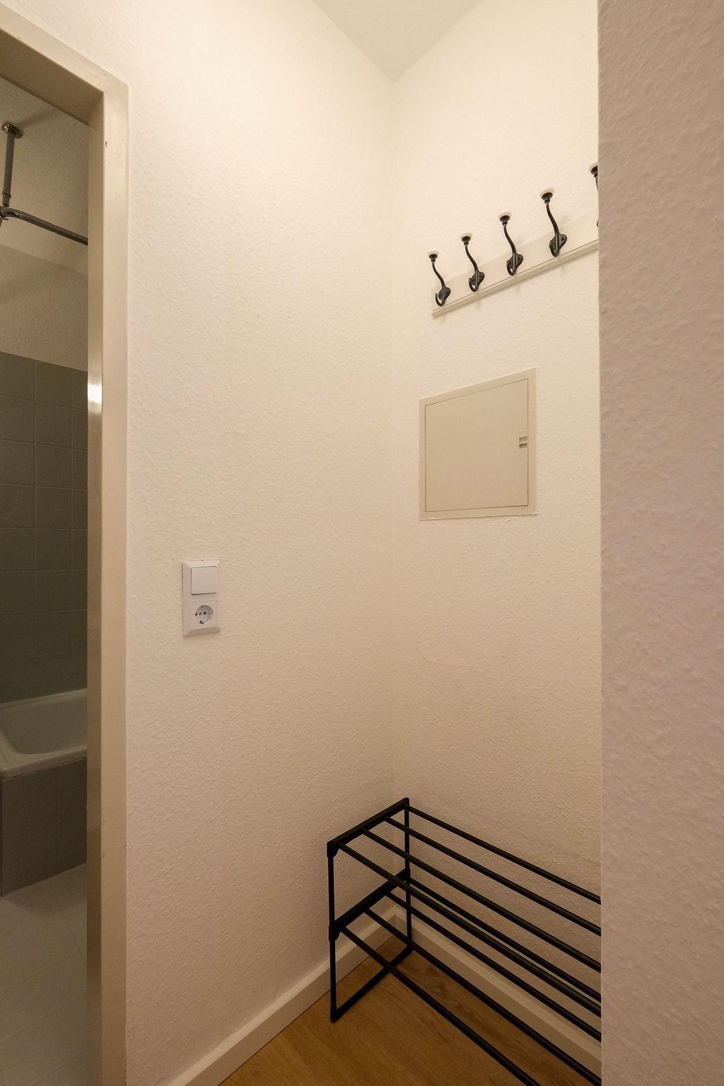 Eine kleine Ecke eines Flurs mit einer schwarzen Metall-Garderobenbank, vier schwarzen Wandhaken und einer geöffneten Tür, die in ein Badezimmer mit grauen Fliesen führt.