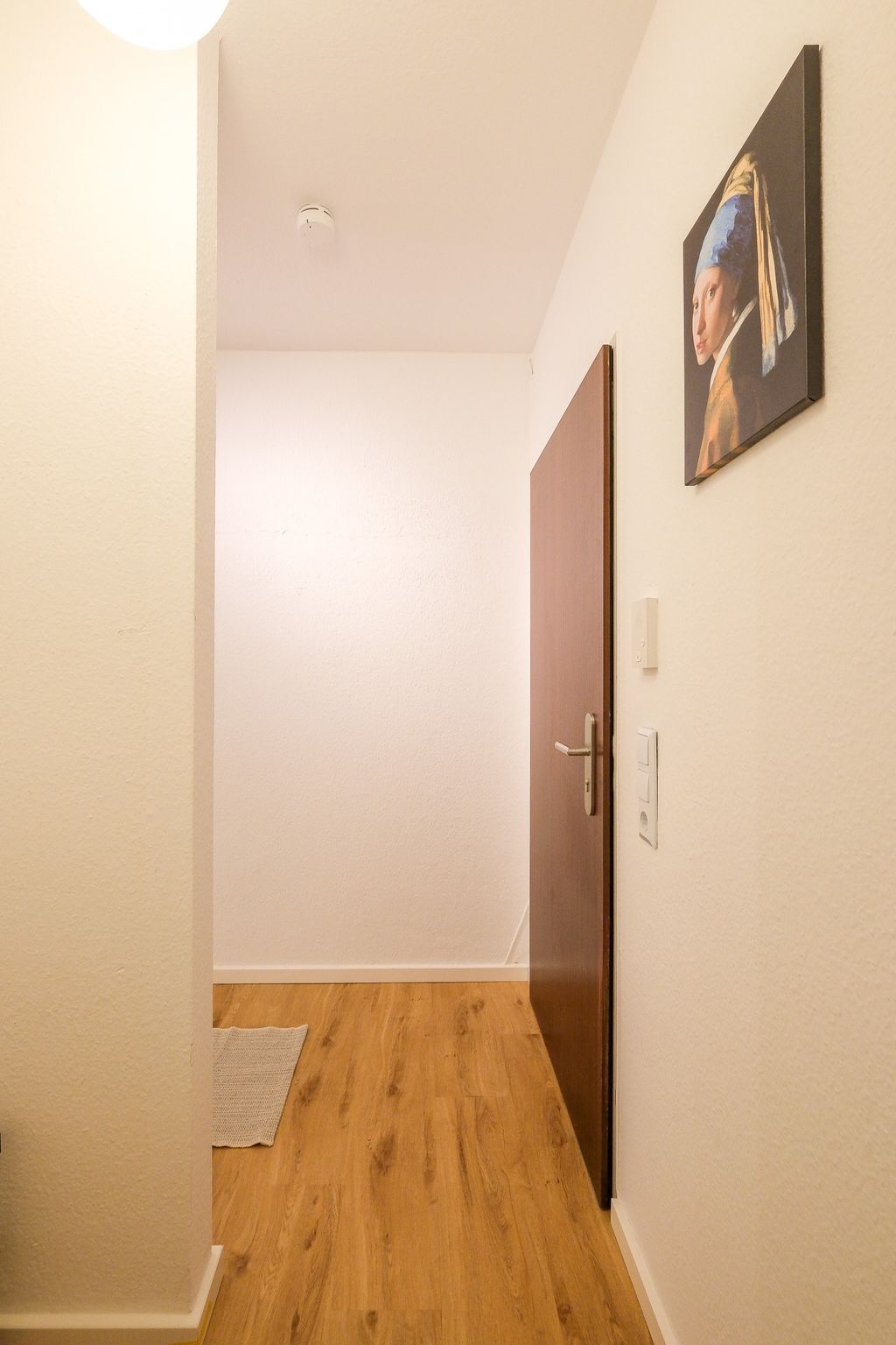 Ein heller Flur mit einer offenen Holztür, einem weißen Wandschalter und einem Gemälde des Mädchens mit dem Perlenohrring an der Wand.