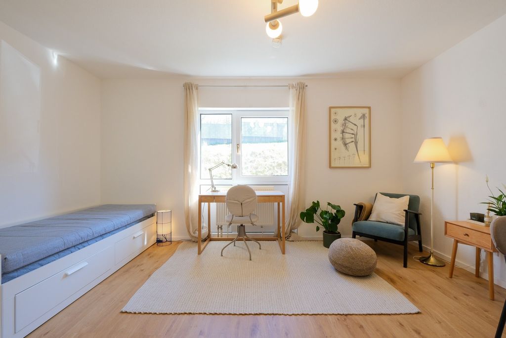 Helles, minimalistisches Arbeitszimmer mit Holzboden, weißem Schreibtisch vor Fenster, grauem Drehstuhl, einem Tagesbett links und einem gemütlichen Sessel mit Stehlampe rechts.