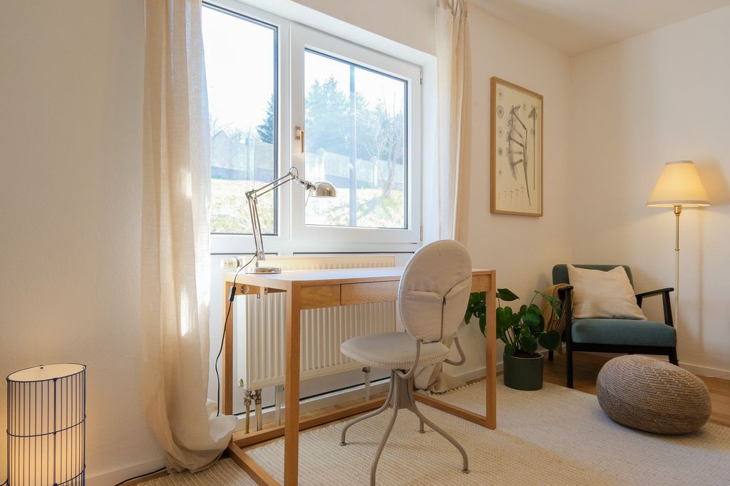 Helles Arbeitszimmer mit einem einfachen Holzschreibtisch vor einem Fenster, einem grauen Bürostuhl, einer grünen Pflanze, einem bequemen Sessel mit Kissen und zwei Stehlampen.