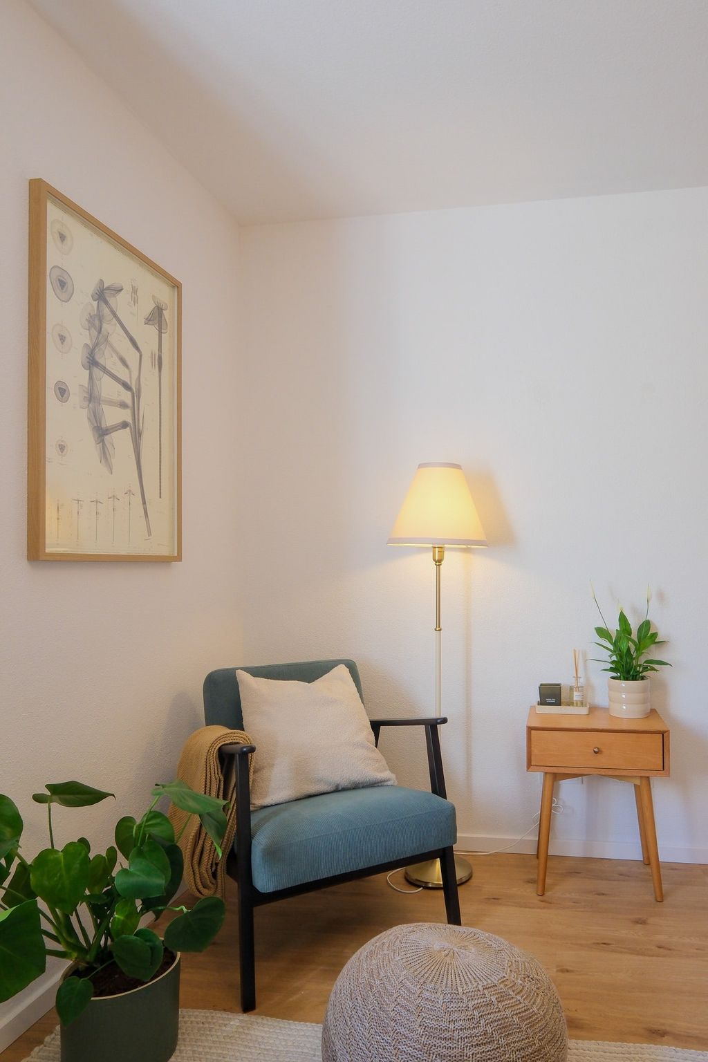 Ein gemütlicher Lesebereich mit einem blauen Sessel, einem Kissen und einer Decke, daneben ein kleiner Holztisch mit einer Pflanze und einem dekorativen Arrangement sowie eine Stehlampe.
