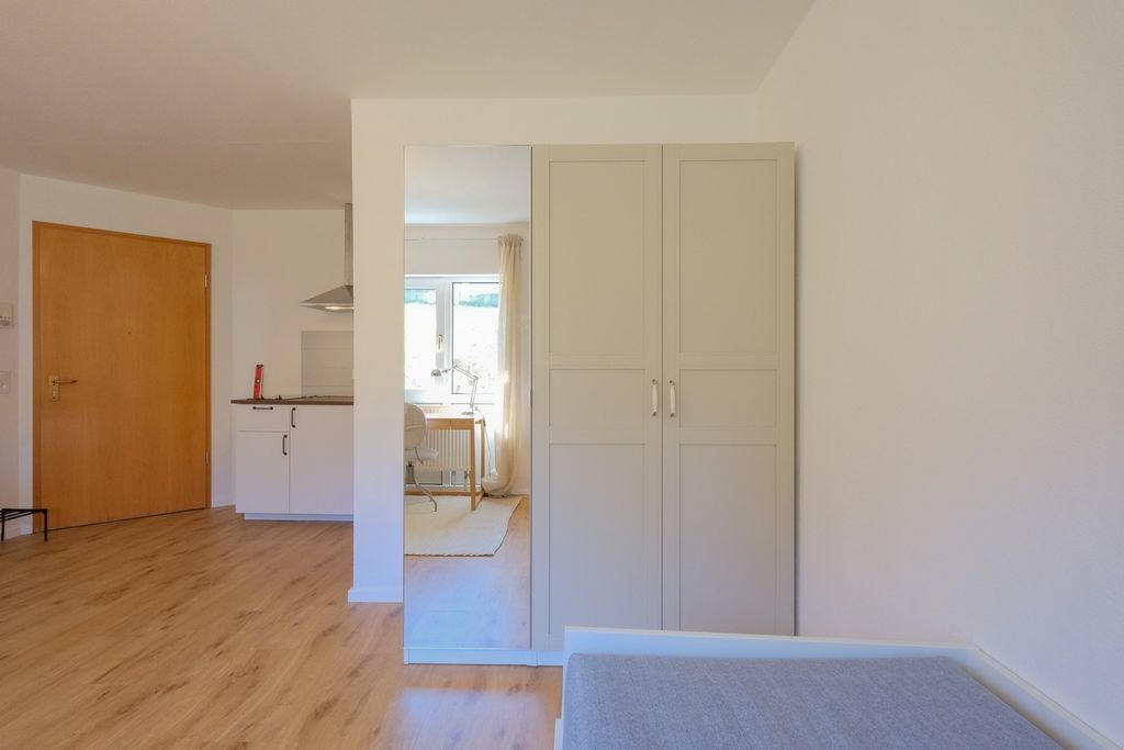 Ein heller Raum mit Holzfußboden, einer weißen Garderobenschrank-Kombination mit Spiegel und einem Teilsicht auf eine Küche und einen Holztürbereich.